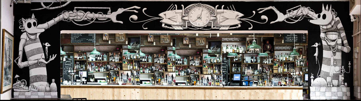 A bar with an extensive selection of drinks is framed by whimsical monochrome murals of cartoon figures with elongated arms and bird companions. The scene has a playful and eclectic atmosphere.